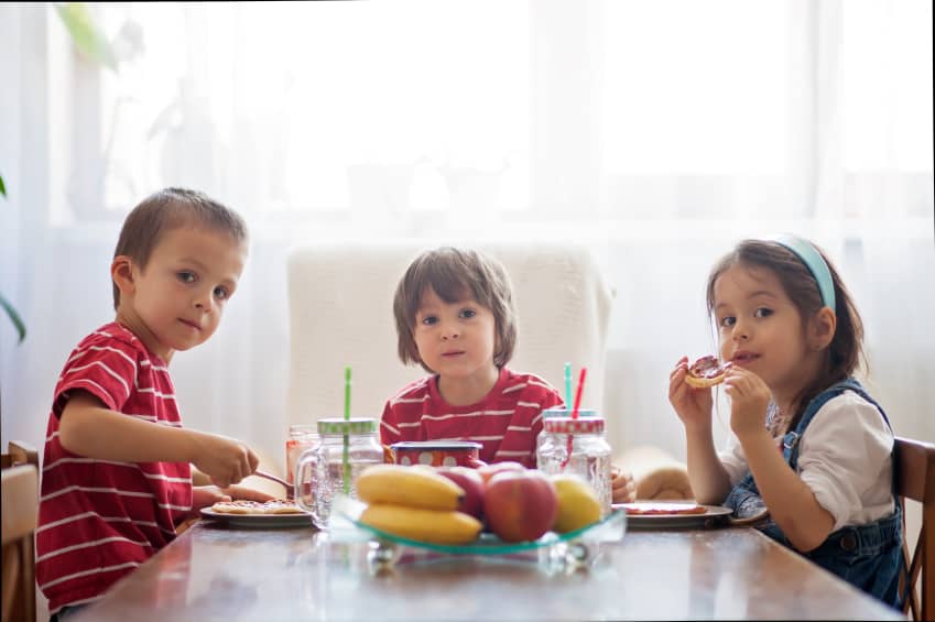 Three happy kids, two brothers and little sister, having healthy breakfast sitting at wooden table in sunny kitchen