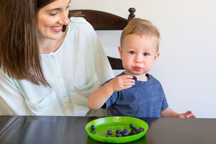 mom and toddler together at the table. Mom watching toddler grasp blueberries as he feeds himself.