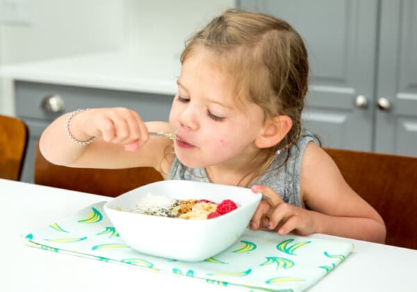 toddler holding own spoon over a bowl of oatmeal and fruit, feeding self