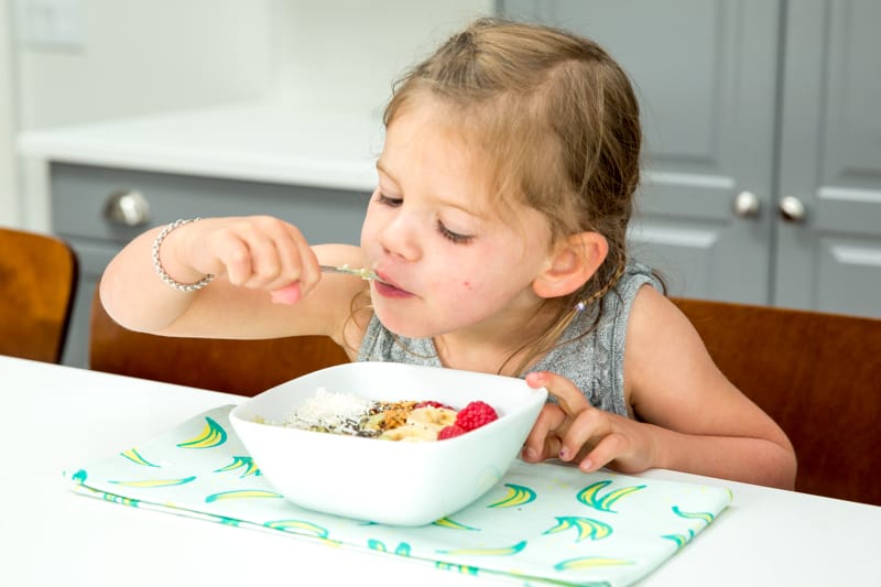 toddler holding spoon feeding self over a bowl of oatmeal and fruit