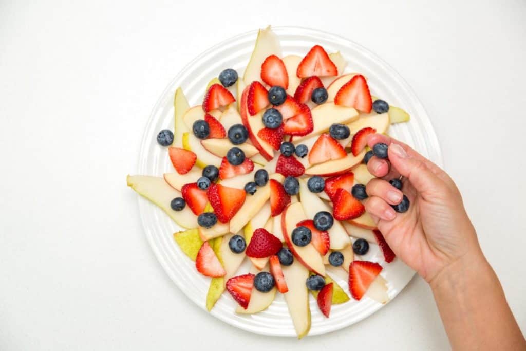 fruit nachos arranged on a white plate with a hand sprinkling berries on them
