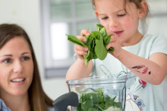 child putting spinach in blender to make green monster blender muffins while mom supervises