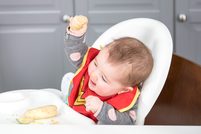 baby in a high chair holding up a baby banana muffin