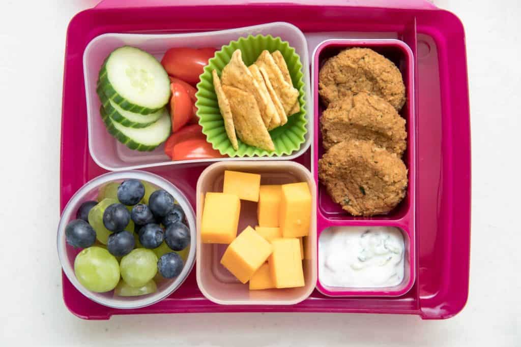 Mittagessen im Bento-Box-Stil mit einer Auswahl an Obst, Käsewürfeln, Crackern, Gemüse und Linsenbissen mit Dip.