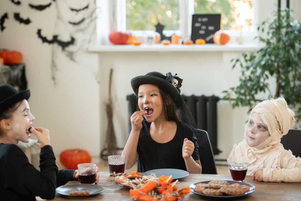 Group of happy kids in Halloween costumes sitting at table in decorated living room and eating cookies and candies at tea party