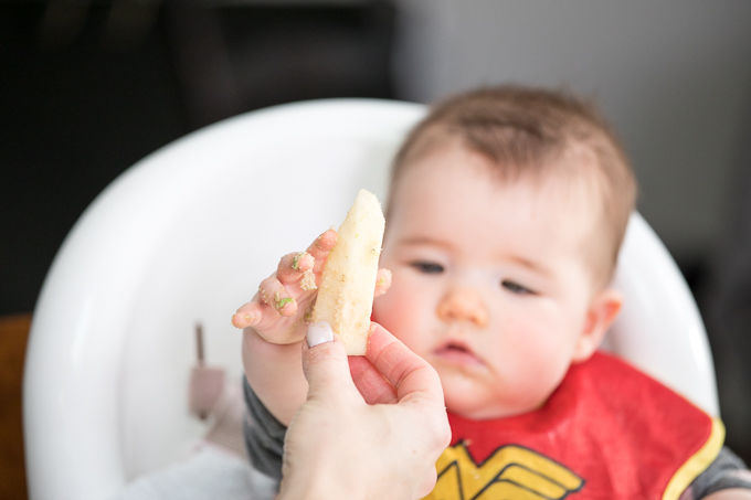 Baby in a high chair reaching for soft finger food held up by adult.