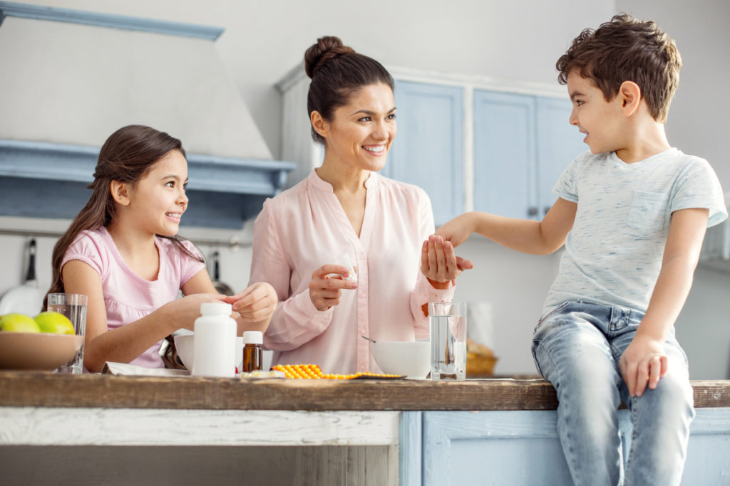 Happy mother. Attractive alert dark-haired young mother smiling and giving pills to her son sitting on the table and her daughter having breakfast