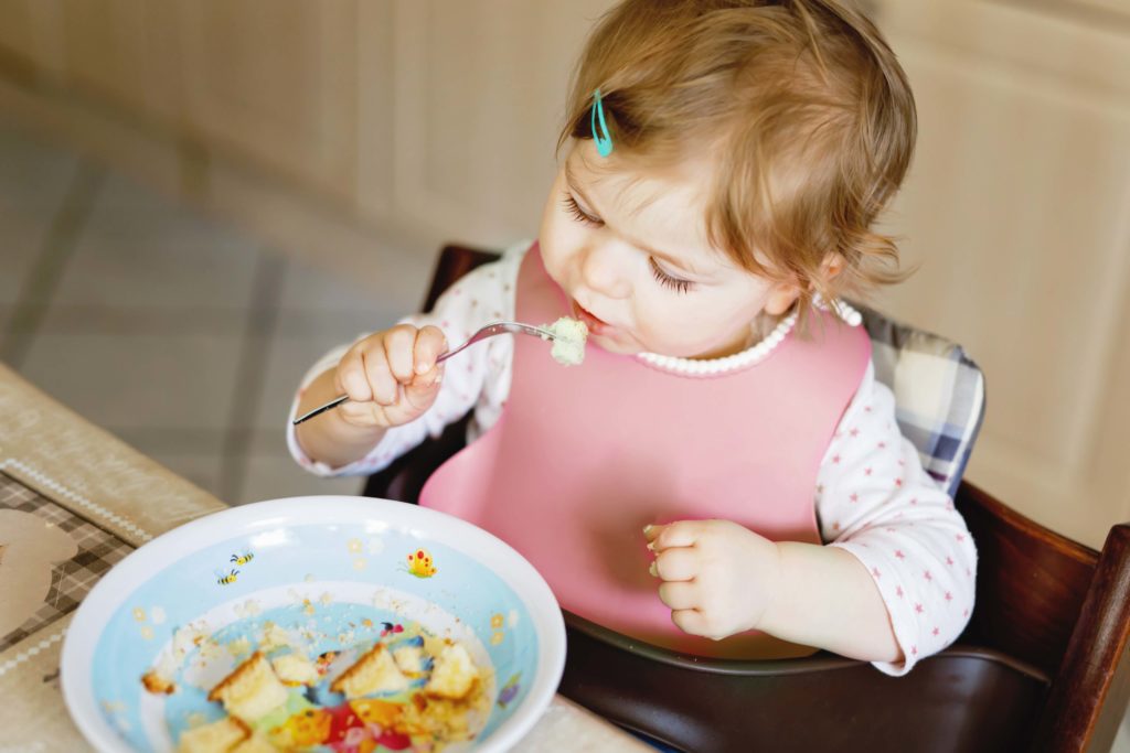 Adorable baby girl eating from spoon mashed vegetables and puree. food, child, feeding and development concept -cute toddler, daughter with spoon sitting in highchair and learning to eat by itself