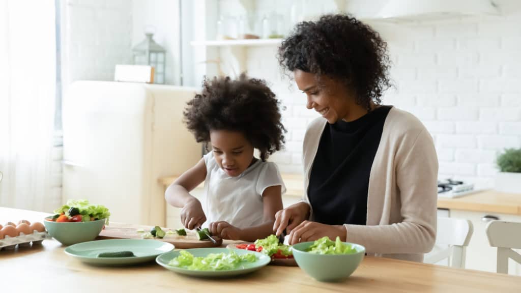Smiling loving African American mother and little daughter prepare healthy delicious salad in kitchen.