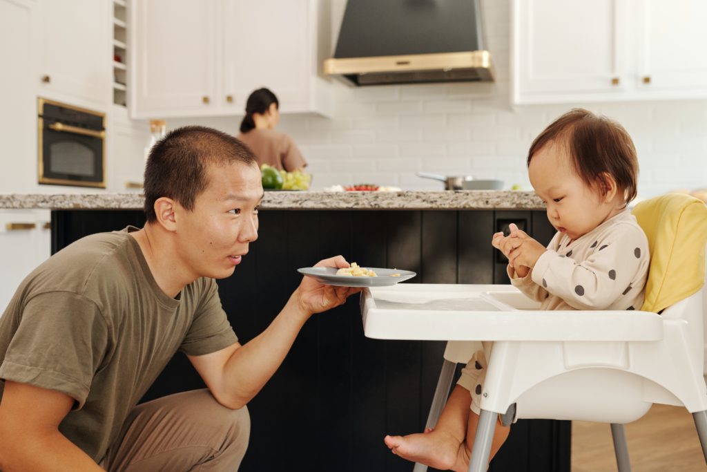 baby in a high chair about to feed herself as dad hands over a plate of food