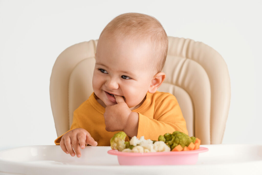 baby sitting in a high chair with a bowl full of soft cooked cauliflower, broccoli and carrots