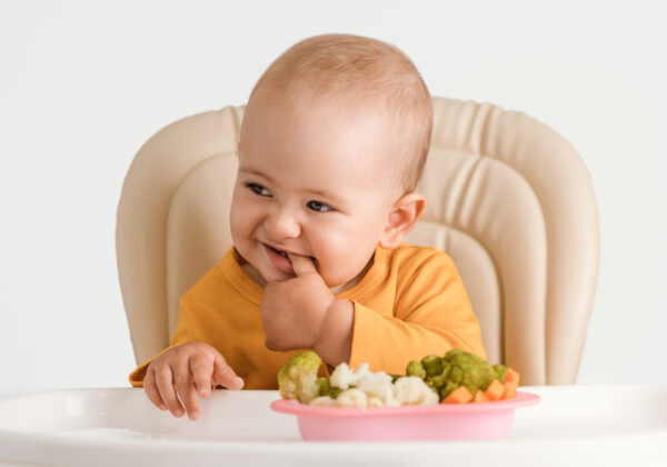 baby sitting in a high chair with a bowl full of soft cooked cauliflower, broccoli and carrots