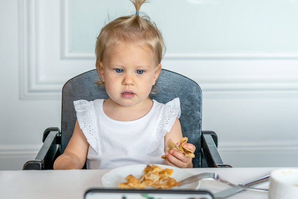 A white Caucasian girl thoughtfully eats pancakes in a restaurant sitting on a child's chair