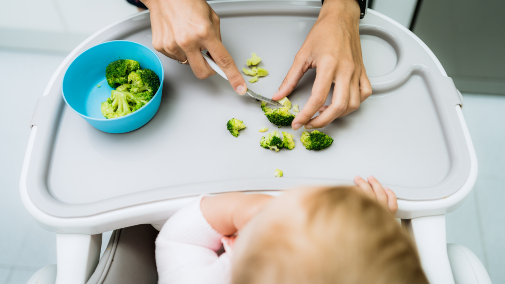 a mother smashing soft broccoli pieces on a highchair for baby-led weaning