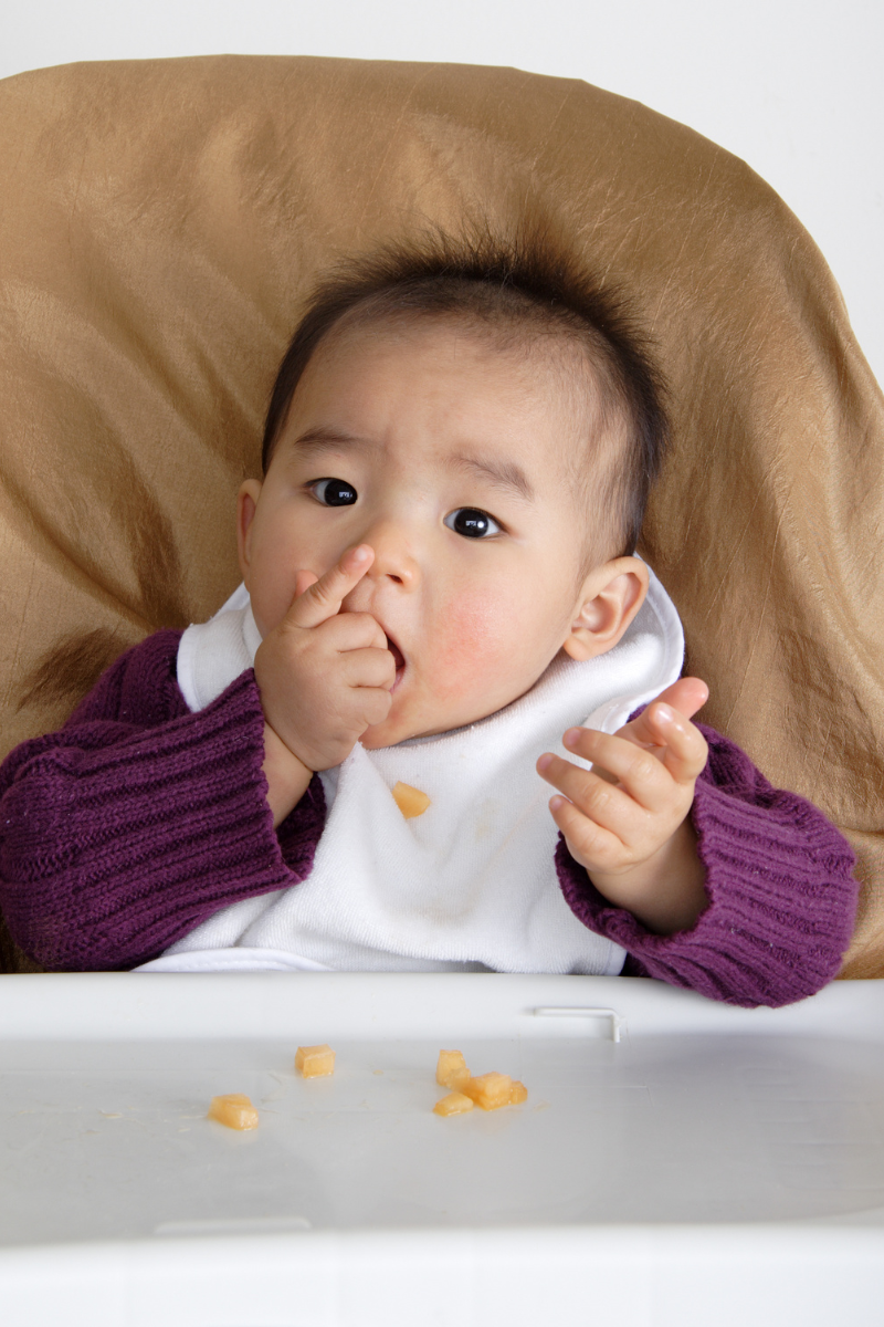 close up of a baby feeding himself pieces of food in a highchair