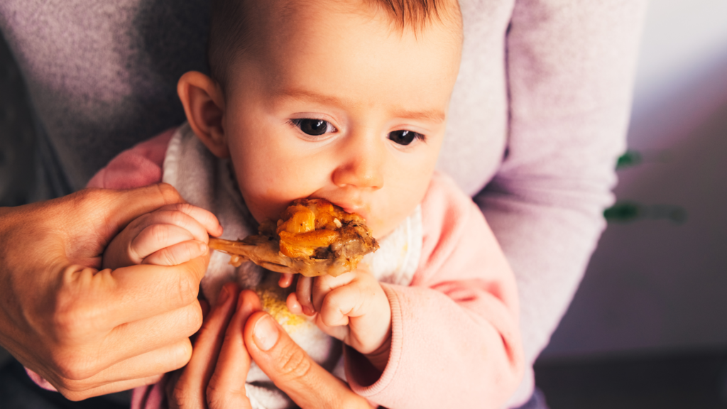 close up of a baby girl holding a chicken drumstick with the help of a parent's hand