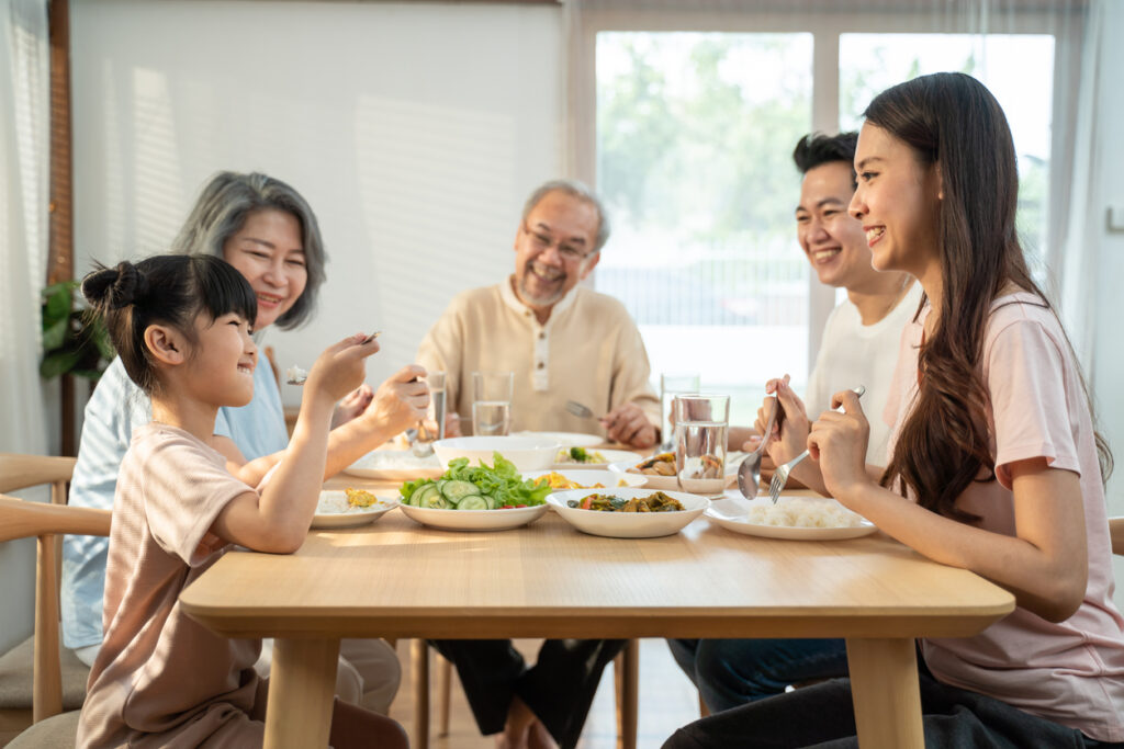 a happy Asian family sitting together at a table. Everyone is smiling at the little girl at the table as she eats