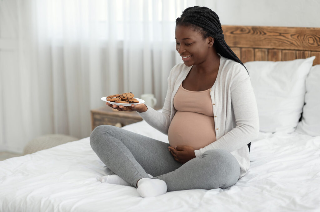 Portrait of black pregnant woman with plate of cookies sitting on bed at home