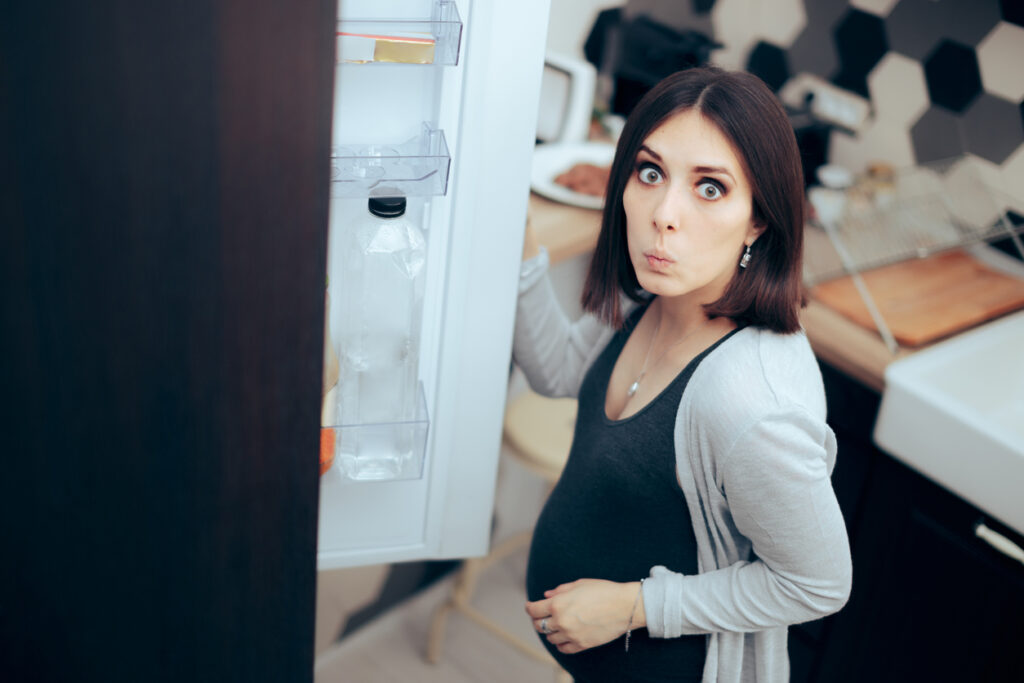 a pregnant woman opening the door to a fridge, looking caught off guard