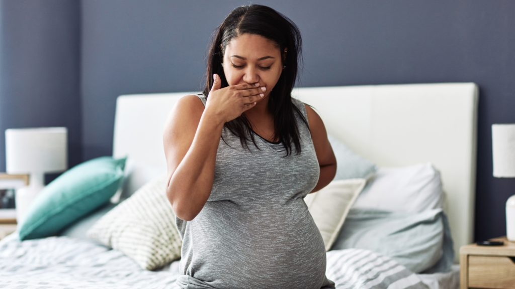 close up of a pregnant women holding her mouth as she experiences nausea on her bed