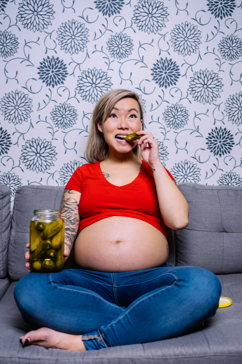 a pregnant woman sitting on a couch holding a jar of pickles