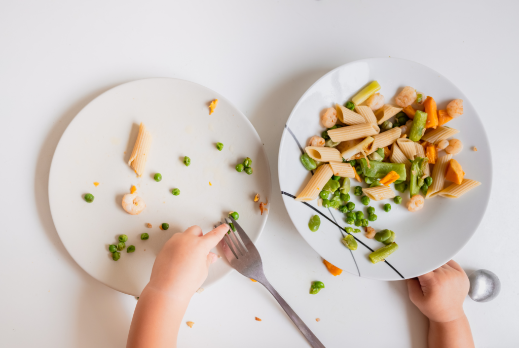 top down view of a child picking away at a plate of food including pasta, peas and shrimp
