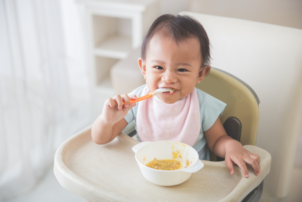 close up of a baby boy feeding himself solid foods in a high chair happily
