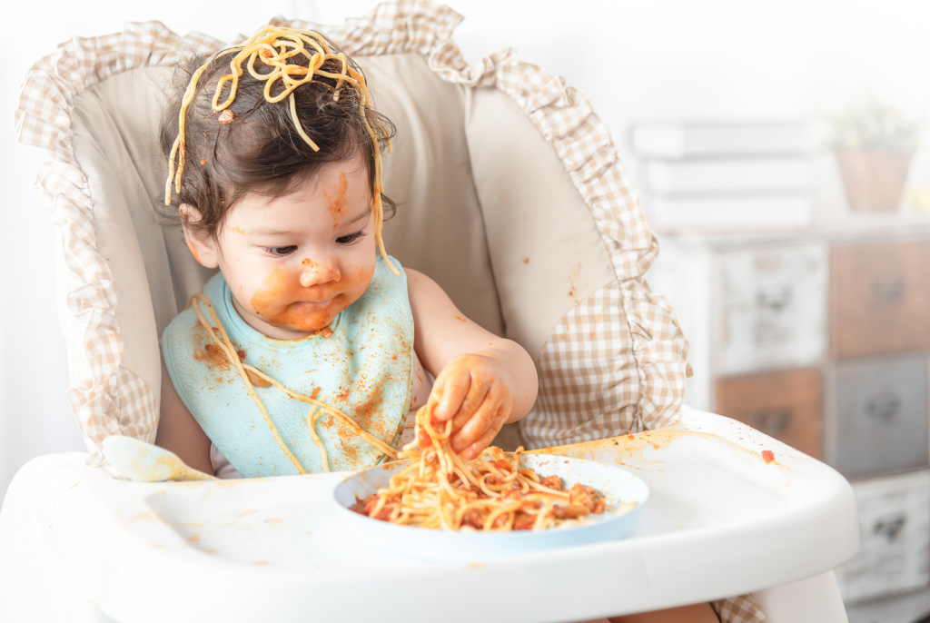 A cute baby girl overwhelmed by the food on her plate in her high chair. She throws spaghetti everywhere.