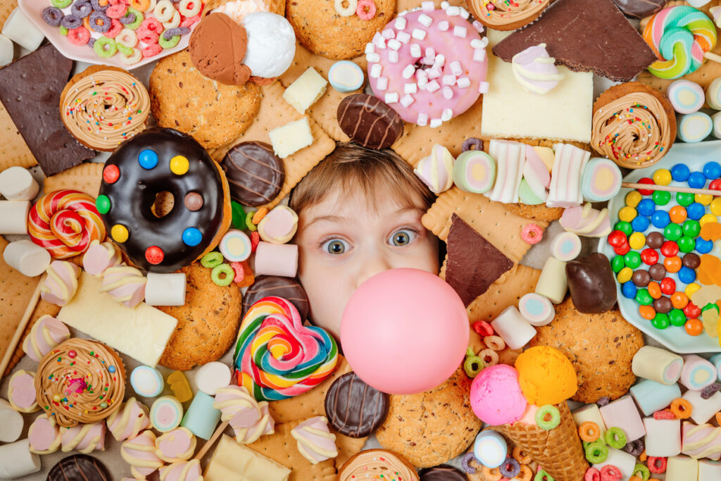 Happy 5 years old girl lying under plenty of sweet foods with bubble gum. Kids face surrounded by unhealthy food. Top view, flat lay.